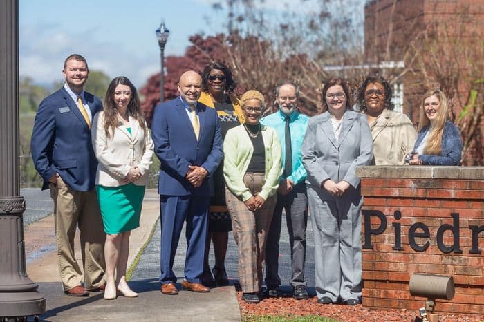 Diverse group of nine professionals smiling outdoors in front of a brick sign.