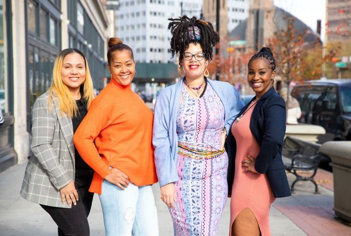 Four smiling women of color stand together outdoors in an urban setting.