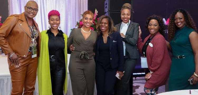 Seven professional women of color smiling and posing together at an indoor event.