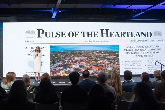 Speaker on stage with large screen displaying 'Pulse of the Heartland' and aerial city view.