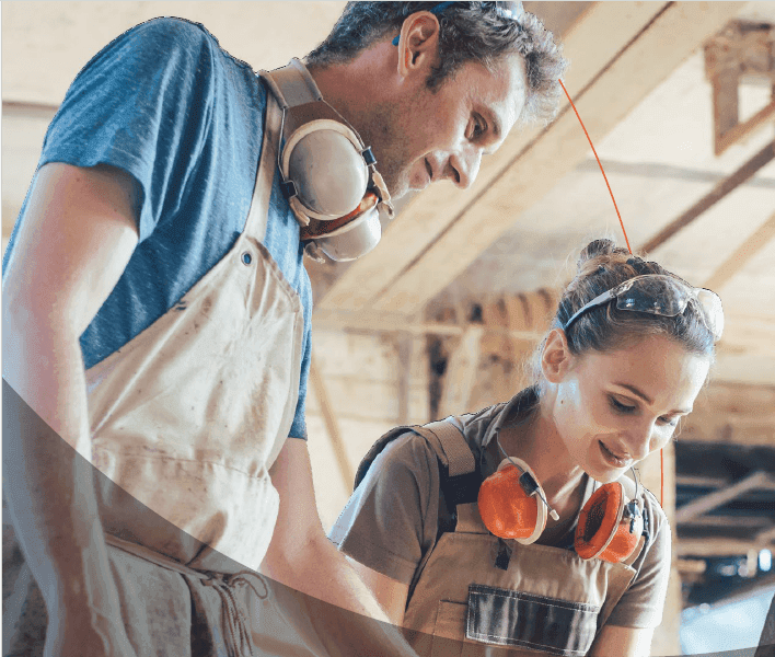 A man and woman wearing safety gear work together in a bright wood workshop.