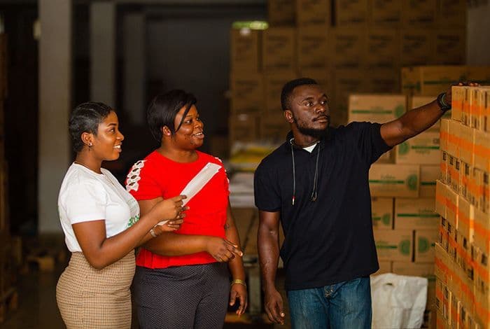 Three workers, two women and a man, inspecting stacked inventory boxes in a warehouse.