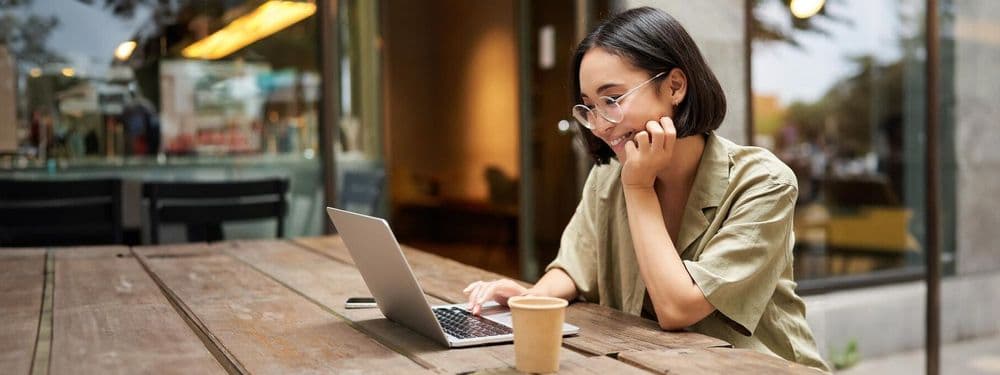 Young woman in glasses works on laptop at an outdoor cafe table, contemplating.
