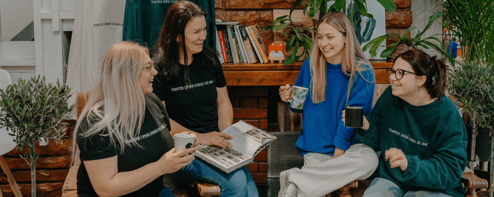 Four women smile and chat casually in a cozy lounge, two holding mugs, one with a magazine.