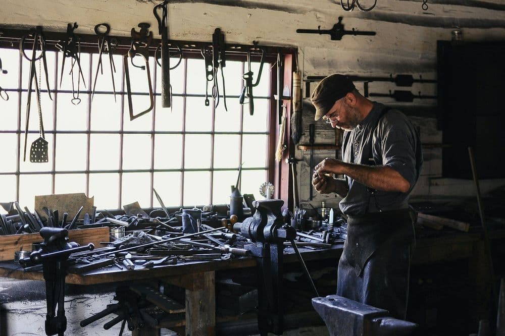 A craftsman meticulously works at a cluttered workbench in a sunlit, rustic workshop.