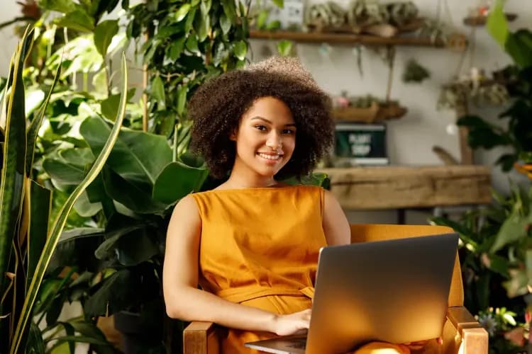 A smiling woman with curly hair works on a laptop amidst lush green plants.