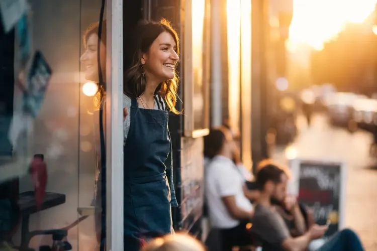 Smiling woman in apron stands in a sunny doorway, looking out at a city street.