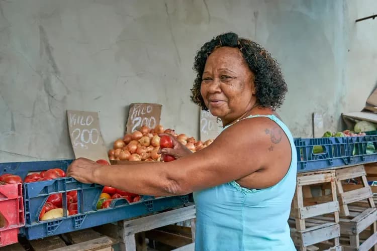 An elderly woman smiles while arranging fresh tomatoes and onions at a vibrant market stall.