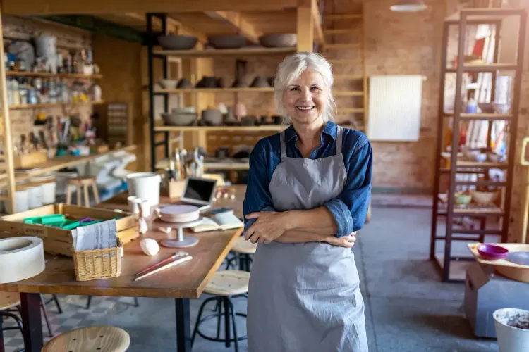 A smiling senior woman in an apron stands in her pottery studio.