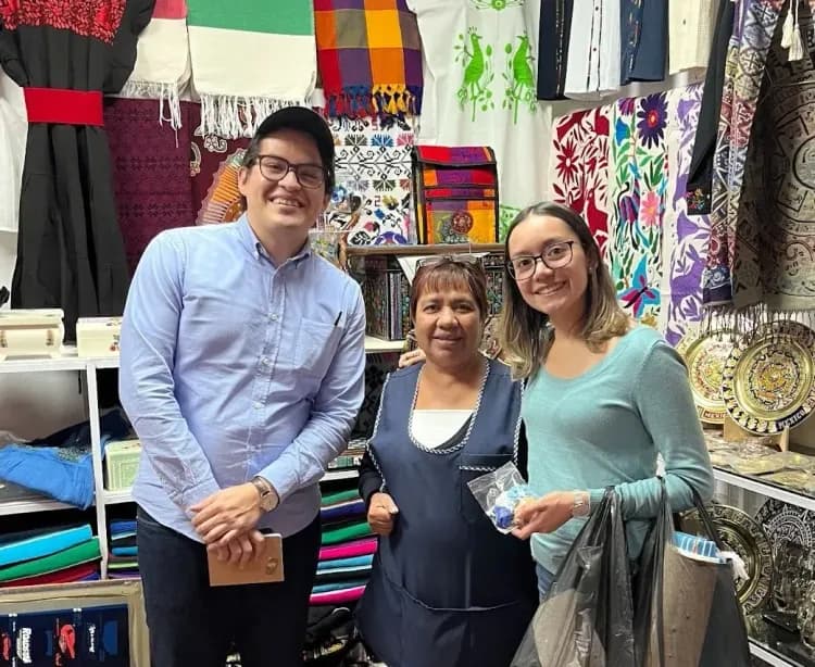 Three smiling people, including a shop owner, pose in a vibrant market stall with colorful textiles.