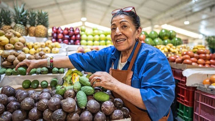 A woman in a blue shirt selling avocados and other fruits at an indoor market.
