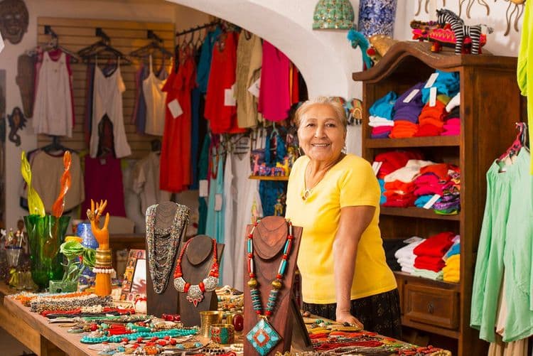 Smiling woman proudly stands in her vibrant craft shop filled with colorful jewelry and clothing.