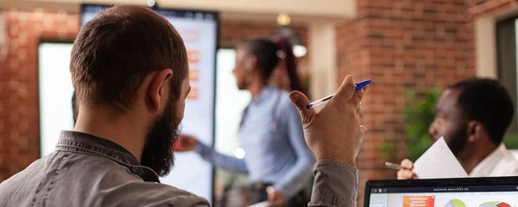 Bearded man watching a woman present on a large screen during a business meeting.
